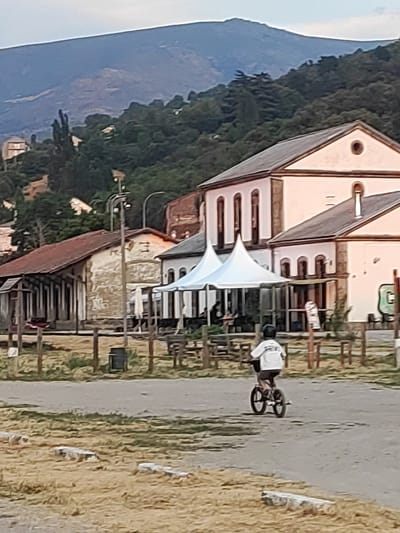 The image depicts a scene in Béjar, Spain, featuring a cyclist in the foreground, a row of buildings, and a mountainous landscape in the background. A young person, wearing a white t-shirt and dark shorts, is riding a small bicycle on a dirt path. The cyclist is facing away from the camera, and is wearing a helmet.
Behind the cyclist are several structures. To the left, there is a long, single-story building with a red-tiled roof and open-air sections. In the center, a large, light-colored building with a dark roof dominates the middle ground. Two white peaked tents are set up in front of this building. The building has arched windows and decorative elements. To the right, another building, also light-colored with a dark roof, is visible.
In the background, a densely forested hillside rises, transitioning into a hazy, mountainous terrain under a pale sky. The overall environment appears to be rural or semi-rural, possibly a former industrial or train station area given the architecture. The ground in the foreground is a mixture of dry grass and dirt.
