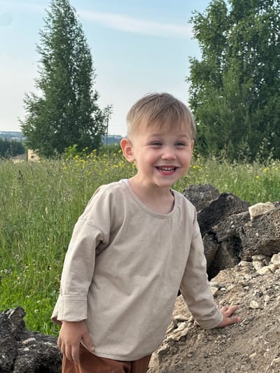 A young boy with blond hair is smiling and looking to his left in front of a pile of rocks and a grassy field. He is wearing a light beige long-sleeved shirt and brown pants. In the background, there are tall green trees and a clear sky. The location is the city of Estachi, Russia.