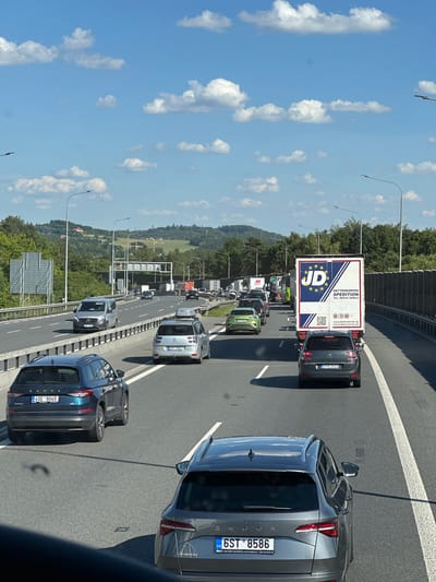 This media file captures a snapshot of a traffic jam on a highway, likely in Mirošovice, Czechia. The perspective is from inside a vehicle, looking forward and slightly upwards. The scene is dominated by a line of cars and trucks, indicating slow-moving or stopped traffic.
The primary subjects are the vehicles themselves, including several cars, a large semi-trailer truck, and other commercial vehicles. The truck in the foreground displays prominent branding: "JD" with stars, and "DETTENDORFER SPEDITION" with contact information and a website. License plates are visible on some vehicles, with one reading "BY 538AX" and another "6ST 8586".
The setting is a multi-lane highway, flanked by a sound barrier on the left and lush green trees and rolling hills on the right. The sky is a bright blue, dotted with fluffy white clouds, suggesting a clear, sunny day. The time of day appears to be daytime, given the bright sunlight casting shadows.
The overall impression is one of a common, albeit frustrating, travel experience. There are no discernible people in focus, and the focus is entirely on the road and the vehicles. The activity taking place is a traffic jam. Notable details include the specific text on the truck, the license plate numbers, and the natural landscape bordering the highway. The mood conveyed is neutral, simply documenting the traffic situation.
