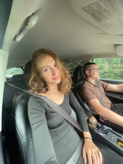 In the city of Sepych, Russia, a man and a woman are seated in the front seats of a car, traveling on a road surrounded by lush green trees. The woman, positioned on the left, is closer to the camera and is wearing a black long-sleeved dress and a seatbelt. She has blonde hair and is looking at the camera with a slight smile. The man, on the right, is wearing a brown short-sleeved shirt and also has a seatbelt on. He is looking forward, presumably driving. The car's interior is visible, including the dashboard and the center console. The lighting suggests it is daytime.