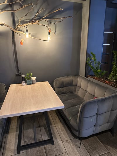 The image shows a cozy dining area within a building in Kirishi, Russia. A light-colored wooden table with black metal legs is positioned in the foreground. On the table, there is a salt shaker, a small potted plant, and a napkin holder. To the right of the table, a plush, gray upholstered couch with tufted detailing is visible. Behind the table and couch, the wall is painted a dark gray. A decorative tree branch with three light bulbs and two small decorative elements hangs from the ceiling. In the background, to the right, there is a window or doorway with reflections and glimpses of green plants in planters. The floor is covered with wood-look tiles. The overall ambiance is warm and inviting, suggesting a restaurant or cafe setting.