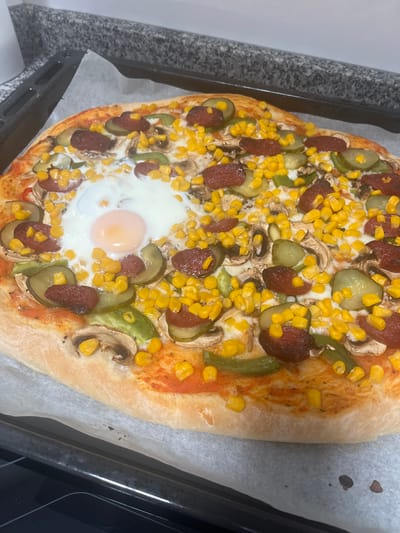 The image shows a freshly baked pizza on a baking sheet. The pizza has a thick crust and is topped with tomato sauce, cheese, corn, sausage, pickled cucumbers, mushrooms, and a fried egg in the center. The baking sheet is lined with parchment paper and is resting on what appears to be a kitchen counter. The image does not provide any information to verify the location as the city of Mogán, Spain.