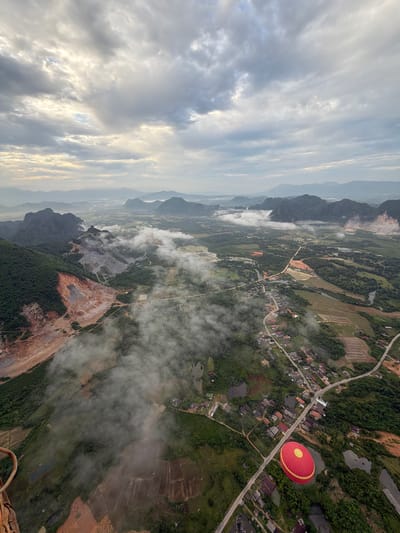 This aerial photograph captures a landscape view of Vang Vieng, Laos, on a cloudy day. In the foreground, a red and yellow hot air balloon is visible, positioned over a rural area with houses and agricultural fields. A paved road winds through the scene, connecting scattered buildings and rice paddies. The middle ground features a lush green landscape interspersed with water bodies and more houses. In the background, dramatic karst mountains rise, some shrouded in mist. A significant portion of the image is dominated by a large, terraced mine or quarry, characterized by exposed rock faces and barren soil, with a landslide visible on one slope. Wisps of fog or low-lying clouds drift across the landscape, adding a sense of depth and atmosphere.