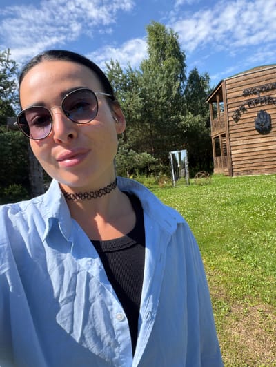 In the city of Rushinovo, Russia, a woman with dark hair and sunglasses is taking a selfie outdoors. She is wearing a light blue open button-up shirt over a black top and a black choker necklace. She is standing on a grassy area with small white flowers. Behind her is a two-story wooden building with a balcony and a sign on the side. To the left of the building, there is a tall mirror placed vertically in the grass, reflecting the trees and sky. Several wire sculptures are also visible in the grassy field. The sky is blue with scattered clouds, and there are trees in the background.