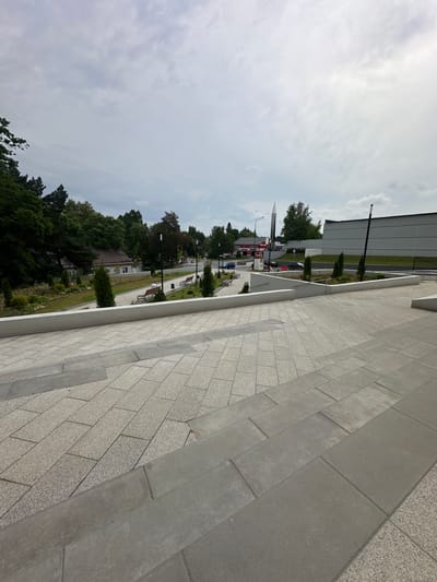 A wide, outdoor, ground-level, eye-level shot shows a paved walkway with large, light grey concrete slabs and smaller, patterned grey stone tiles. The walkway leads up to a white, low-lying wall and then to a park-like area. The park has green grass, trees, bushes, and several park benches. In the background, a modern, silver, rocket-shaped monument stands tall against a cloudy, pale blue sky. To the left of the monument, a building with a red roof and white walls is visible, with parked cars in front of it. To the right of the monument, a large, grey, industrial-looking building with a dark grey roof dominates the scene. A tall, thin, black and white pole stands in front of this building, and another similar pole is visible further to the right. The location is identified as Vlasikha, Russia, a city known for its military aerospace defense installations.