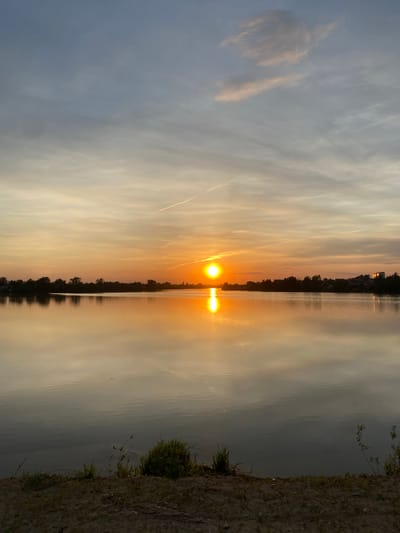 The image depicts a sunset over a body of water, likely a lake or wide river. The sun is centered, with its reflection extending vertically downward in the water. The sky is a mix of pale blue and orange, with wispy clouds streaking horizontally. The water is calm, reflecting the colors of the sky. Two strips of land, likely shorelines, run along the left and right sides of the water, with sparse vegetation visible. There are buildings in the distance. Based on the location requested, it is possible the scene is near Aramil, Russia, but there is nothing within the image itself to confirm the exact location. There are no people, actions, or interactions visible in this still scene.