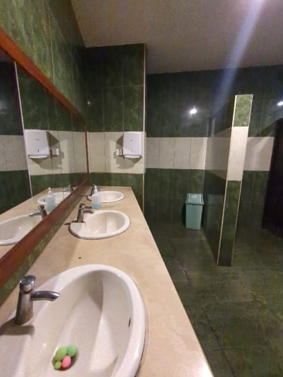 The image depicts a bathroom sink located in a public restroom in Morogoro, Tanzania, at the coordinates -6.835 latitude and 37.672 longitude. There are four white porcelain sinks, two of which are visible, with one in the foreground, containing three brightly colored balls. The sinks are mounted on a counter with a beige-colored surface, with green tiled walls surrounding the counter. The bathroom has a large, rectangular mirror above the sinks. The floor is tiled with green and beige tiles, while a green trash bin is visible in the background. The bathroom is clean and well-lit, with a white ceiling and a white door. There is a hand dryer on the wall and two bottles of hand sanitizer on the counter.