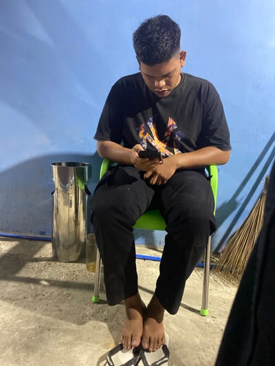 The image depicts an individual, likely male, seated on a bright green plastic chair within what appears to be a casual indoor or semi-enclosed space in Gampongaceh, Indonesia. The person has dark hair and skin, and is dressed in a black t-shirt with a vibrant, abstract graphic design, paired with black pants. His gaze is directed downwards, focused intently on a black smartphone held in both hands, indicating deep engagement with the device.

The setting features a plain blue-painted wall in the background, against which a broom made of natural fibers leans. The floor is made of concrete. To the left of the chair, a large, reflective silver cylindrical object, possibly a thermos or water dispenser, rests on the floor. A blue pipe or conduit runs horizontally along the base of the wall. The person's feet are bare, with one white and black sandal, distinctly branded "SWALLOW" on its strap, visible near his feet.

The lighting suggests a nighttime or dimly lit indoor environment, with a strong light source creating prominent shadows on the floor and wall. The scene captures a moment of quiet absorption in a digital activity within an unpretentious, everyday setting.