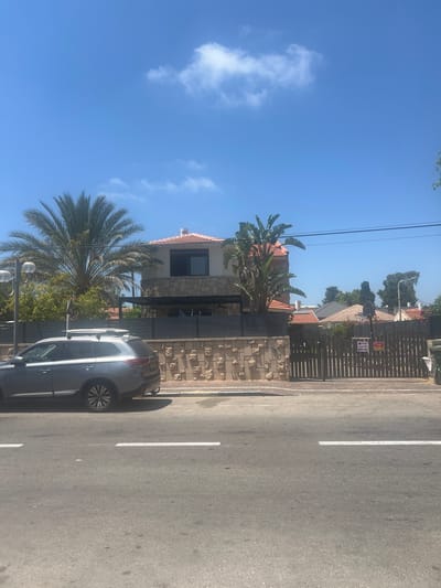 This image depicts a residential street scene in the city of Or Akiva, Israel, under a bright blue sky with sparse white clouds. In the foreground, a gray SUV is parked on the side of an asphalt road with a white line marking. Behind the car, a light brown stone wall with decorative patterns runs along the property line. A dark wooden fence with vertical planks encloses the property. A sign with text in Hebrew is attached to the fence. Beyond the fence, a house with a reddish-orange tiled roof is visible, featuring stone facade details and a dark-framed window on the upper level. Several palm trees and other foliage are present on the property. The environment appears to be a sunny day in a suburban area.