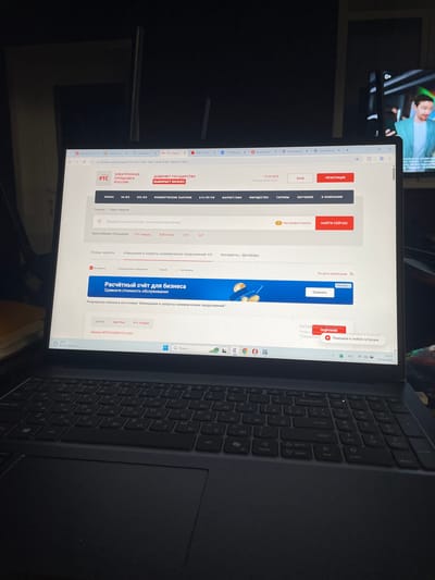 A forensic examination of the image reveals an indoor scene in Chulym, Russia, depicting an open, black HP laptop positioned on a dark surface, likely a desk or table. The laptop screen displays a web browser (Microsoft Edge) actively viewing the Russian procurement website rts-tender.ru, specifically a search results page for "Закупка №32616080519 в ЕИС" (Procurement №32616080519 in EIS). The website's displayed time is "19:33 МСК" (Moscow Standard Time) on "Пятница 05.06.2026 г." (Friday 05.06.2026). The laptop's taskbar shows the system time as "23:33 05.06.2026," indicating a 4-hour difference from Moscow Standard Time, consistent with the UTC+7 time zone of Chulym, Russia. The taskbar also displays a weather icon showing "22°C В осн. облачно" (mostly cloudy) and a "РУС" (Russian) language setting. A "Активация Windows" (Windows Activation) pop-up is partially visible on the screen. The laptop features a black QWERTY keyboard with Cyrillic characters and a numeric keypad. In the background, to the right of the laptop, a flat-screen television is displaying a male figure with dark hair, wearing a light blue jacket over a white shirt, smiling and looking left. A "0+" age rating logo is visible in the top right corner of the television screen. The room appears dimly lit. No individuals are present within the immediate scene.