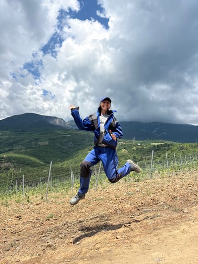 A woman is captured mid-air, jumping with her arms outstretched, in what appears to be a vineyard. She is wearing blue work clothes, a blue cap, and sturdy boots. The background features rolling green hills covered in trees, with a few power lines visible on the slopes. The sky is filled with dramatic, cloudy formations. The ground in the foreground is dry and rocky, with rows of vineyard trellises extending into the distance. The scene suggests a rural, outdoor setting.