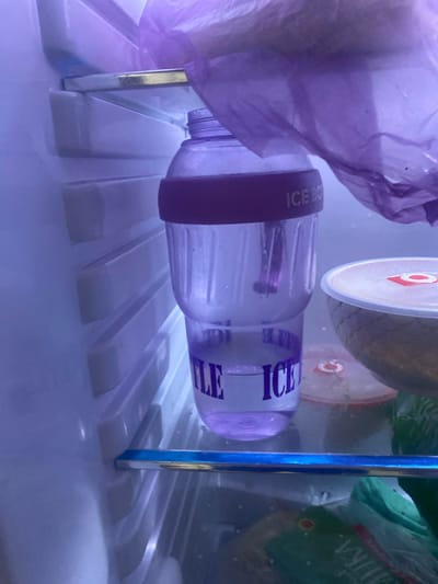 A purple plastic water bottle with "ICE B" printed in white on a purple band is visible in a refrigerator in Kemin, Kyrgyzstan. The bottle is half-filled with a purple liquid and has a purple straw inside. To the right of the bottle, a white bowl with a red and white circular label is partially visible. The refrigerator shelves are white and appear to be made of plastic. A purple plastic bag is draped over the top of the bottle. The overall lighting is dim and has a purple hue.
