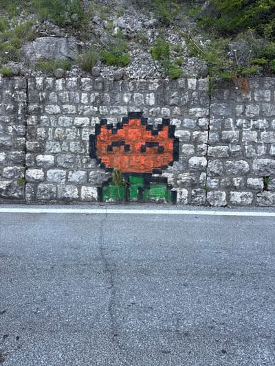 An orange and black pixelated alien-like figure is spray-painted on a grey stone wall. The figure has a square-shaped head with two black dots for eyes and a black outline. Below the head, it has green legs with black detailing. The wall is made of rough-hewn rectangular stones of various shades of grey and is cracked in several places. Above the wall, there is a rocky incline covered with sparse green vegetation and what appears to be a metal mesh fence. A white line marks the edge of a grey asphalt road in the foreground. The scene is located on the side of a road in Bukovik, Montenegro.