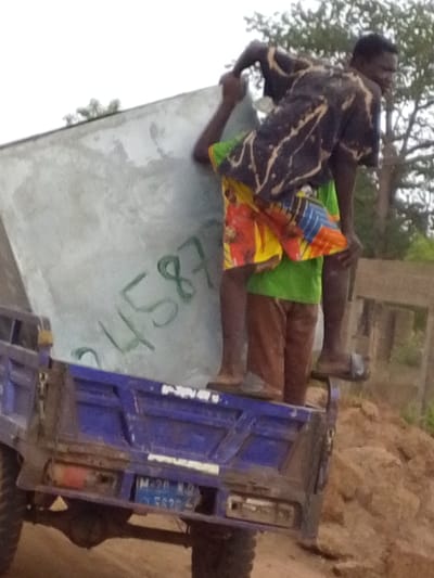 In Dungu, Ghana, two men stand on the back of a blue truck. One man is holding a large, grey rectangular object with the number "24587" written on it. The other man is standing behind him, also holding the object. The truck is parked on a dirt road, with trees in the background.