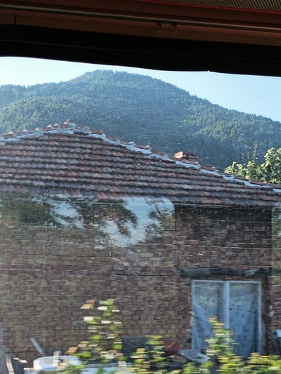 The image shows a view through a window, likely from a moving vehicle, of a brick building with a tiled roof. In the background, a dense, tree-covered hill rises against a clear blue sky. The building's roof is made of dark, weathered tiles, some of which appear to be cracked or missing. The walls are constructed from red brick. A reflection on the window obscures parts of the building's facade, showing distorted images of trees and the sky. To the right of the building, a portion of lush green foliage is visible. On the lower left side of the image, out-of-focus green plants are visible, along with what appears to be a white basin or container. Two doors with frosted glass panels are visible on the right side of the building's facade. The overall scene suggests a rural or semi-rural setting, possibly in the vicinity of Chelopech, Bulgaria, given the presence of vegetation and a mountainous backdrop. There are no people or animals clearly identifiable in the image.