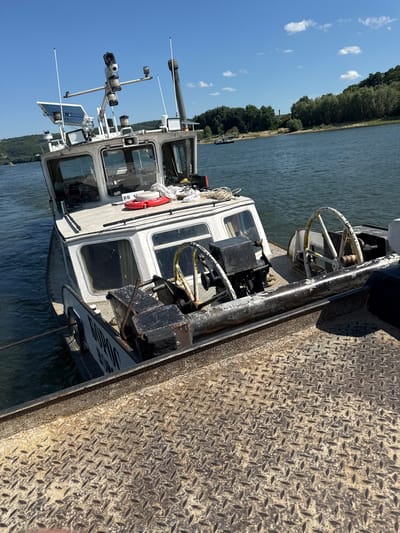 This is a medium-angle, eye-level, outdoor photograph taken on a sunny day, depicting a tugboat docked next to a larger vessel. The tugboat, the primary subject, is white with a dark hull and appears to be operating on a body of water. It is equipped with various navigational and operational equipment, including antennas, lights, solar panels, and what appear to be cameras mounted on a gantry structure. Two ship's wheels are visible on the deck of the tugboat, one of which is partially obscured. The name "GOPHER" is visible in black lettering on the side of the tugboat. The larger vessel, in the foreground, has a metal deck with a diamond plate pattern. The background shows a wooded shoreline with trees and some distant structures. The water is calm with moderate ripples. The scene is consistent with a port or docking area in Cherkovitsa, Bulgaria. There are no people clearly visible in the image.