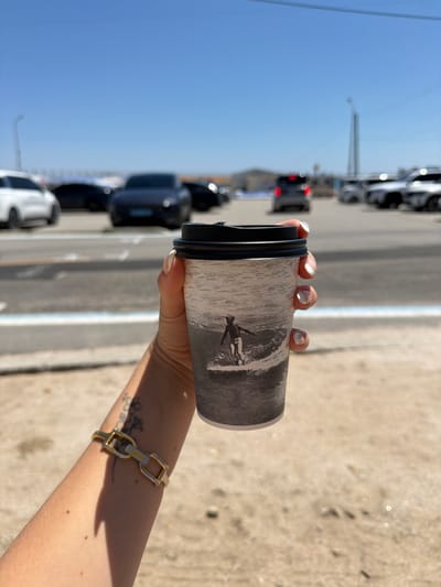 A person's arm extends into the frame, holding a coffee cup. The cup is gray with a black lid and features a monochrome image of a surfer on a wave. The person wears a gold bracelet. The background shows a street with cars parked along the side. The sky is clear and blue. The location appears to be in a populated area with urban characteristics.