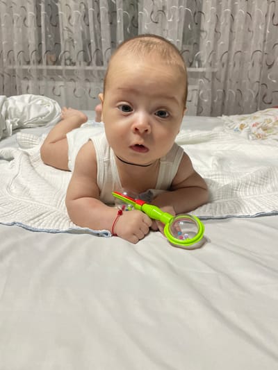 A baby is lying on its stomach on a bed, holding a green rattle. The baby is wearing a white tank top and has a red string bracelet on its left wrist. The bed has white sheets and a white blanket with a blue trim. In the background, there are sheer curtains. The scene appears to be indoors, likely in a bedroom. There is no information in the image that suggests the location is Fălești, Moldova.