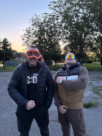 Two adult males stand outdoors on a paved surface, possibly asphalt, with trees and vegetation in the background. The location is identified as Topoli, Bulgaria. The male in the foreground wears a black quilted jacket, black pants, and orange goggles. The second male behind him is wearing a brown vest over a darker jacket, brown pants, and reflective goggles. Both individuals are facing towards the camera. The sky suggests either sunrise or sunset.