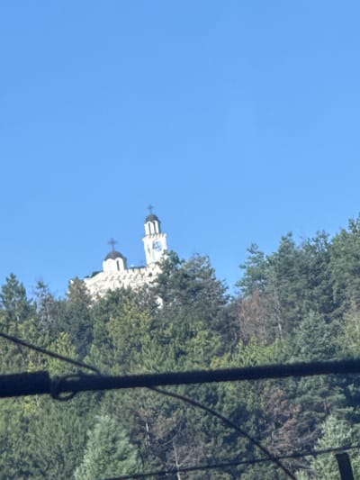 The image captures a panoramic view of the Holy Trinity Church in Hadzhidimovo, Bulgaria, perched atop a verdant hill. The clear blue sky serves as a backdrop to the imposing white church, its architectural grandeur enhanced by two prominent domes, each topped with a cross. A tall bell tower, also white, stands beside the main structure, featuring a clock face and a cross. The hillside is densely populated with a variety of trees, their lush green foliage contrasting with the white of the church. In the foreground, dark, blurred horizontal lines suggest the presence of electrical wires or cables, indicating a possibly urban or semi-urban setting. The overall scene is one of serene elevation, with the church acting as a focal point against the natural backdrop.