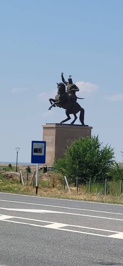 The image shows a bronze equestrian statue of a warrior on a pedestal. The statue is located in an outdoor setting with a clear blue sky and some clouds. In the foreground, there is a road with white markings and a traffic sign indicating a camera. There are also some fences and vegetation around the statue. The scene appears to be in a public area.