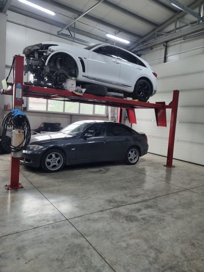 The image shows a two-car automotive repair shop in Pobeda, Bulgaria. A white Infiniti SUV is lifted on a red four-post lift with the front bumper and headlights removed, exposing the engine and front suspension. Next to it, a dark gray BMW is raised on a similar lift. The shop has a concrete floor with expansion joints and fluorescent lighting. There are windows in the background. There are no people visible.