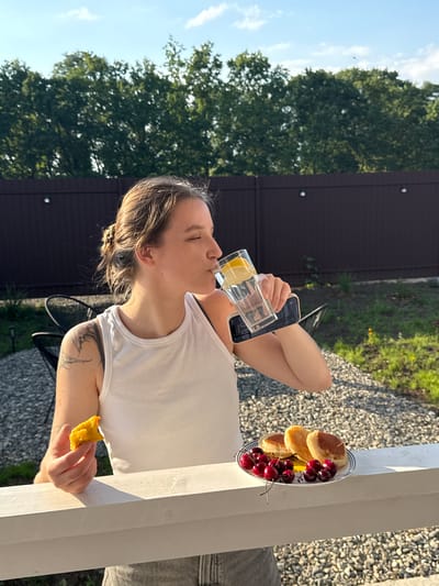 A young woman is pictured outdoors, likely on a porch or patio, in Starokorsunskaya, Russia. She is holding a glass of water with lemon and taking a sip, with a phone resting beneath the glass. In her other hand, she holds a piece of food, possibly a pastry. In front of her, on a white railing, is a plate with three small pancakes and a pile of cherries. The background features a dark brown fence, lush green trees, and a bright blue sky with scattered clouds. The overall setting suggests a warm, sunny day.