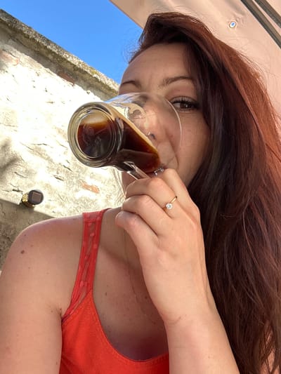 A woman with long, dark brown hair drinks from a glass of dark liquid, possibly coffee or a dark beverage. She is wearing an orange tank top with a textured fabric on the straps. A thin gold necklace is visible around her neck, and she is wearing a gold ring with a single diamond on her left hand. She is seated outdoors, with a stone wall and a clear blue sky in the background. The scene appears to be in Villa Pasquali, Italy. The lighting suggests it is daytime.
