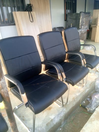The image displays a row of three black, executive-style chairs with chrome frames, positioned on a concrete surface. The chairs appear to be new or in very good condition. Behind the chairs, stacked pieces of light-colored wood are visible, leaning against a wall. To the right of the chairs, several stacked items wrapped in plastic sheeting are present, suggesting they might be for sale or storage. The environment appears to be an outdoor or semi-outdoor area, possibly a warehouse, showroom, or market. The lighting suggests it is daytime. The overall scene is static and devoid of human activity. The context provided about the city of Indo, Nigeria, is not visually derivable from the image itself.