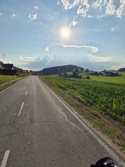 The image depicts a rural road scene in St. Oswald bei Freistadt, Austria. The road is asphalt with white lane markings, showing signs of wear and tear with visible cracks. To the right of the road is a field of lush green corn. In the distance, rolling green hills are covered with trees and dotted with houses. The sky is bright blue with scattered white clouds, and the sun is shining brightly, creating lens flare. The overall impression is a sunny day in the Austrian countryside.