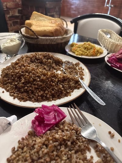 This is a close-up, eye-level shot of a table laden with food, suggesting a meal in Novy Urengoy, Russia. The lighting is warm and somewhat dim, creating a cozy atmosphere. The table's surface is black and glossy, reflecting the dishes.
In the foreground, a white plate contains a generous portion of cooked buckwheat. A fork rests on the plate, its tines digging into the grains. To the side of the buckwheat is a small mound of bright pink pickled cabbage. To the right of this plate, another white plate holds what appears to be pilaf, a rice dish with visible pieces of orange carrot. A small wicker basket with a spoon sits behind the pilaf.
In the mid-ground, a larger white plate is filled with more buckwheat. A silver spoon is resting on this plate. To the left of the buckwheat, a glass gravy boat contains a creamy white sauce, likely sour cream or a related condiment. Behind the gravy boat and buckwheat, a woven basket holds several slices of toasted white bread.
The background shows a brick wall, possibly part of the restaurant's decor, and a glimpse of white seating. The overall impression is a hearty, traditional meal, possibly at a restaurant or a home setting in Novy Urengoy. There are no people visible in the image.