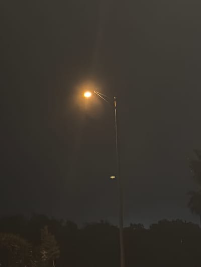 The image depicts a street lamp at night in Englewood, United States. The lamp emits a bright, warm light that illuminates the dark sky around it. Below the lamp, silhouetted trees form a dark line against the horizon, suggesting a park or natural area. The overall atmosphere is serene and peaceful, with the solitary street lamp providing a point of light in the otherwise dark environment.