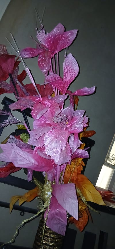 The image depicts a close-up view of a decorative floral arrangement, predominantly in shades of pink and accented with gold and orange foliage. The arrangement features large, artificial flowers with layered, textured petals that have a shimmering, iridescent quality. Some petals are adorned with embossed patterns resembling feather veins. Among the larger blooms are smaller, spiky metallic accents and delicate, translucent leaf-like structures. The arrangement is further embellished with gold, bead-like strings and metallic leaf-shaped elements in orange and yellow hues. The background is a plain, muted gray wall, with a sliver of a window visible on the right side, showing a white blind and a dark, possibly metal, frame. The overall impression is of an indoor decorative piece, likely placed on a surface or in a vase, given the obscured base which appears to be a textured gold container. There is no indication of any specific location within the city of Andijani, Uzbekistan, nor are there any people or actions depicted.