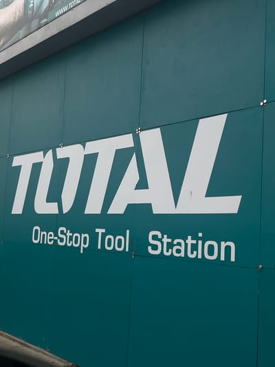 The image shows a close-up of a teal-colored building facade with large white lettering. The prominent text reads "TOTAL" in a bold, sans-serif font. Below "TOTAL", smaller white text states "One-Stop Tool Station". Several small circular metallic fixtures are visible, evenly spaced along the horizontal and vertical seams of the facade panels. The building appears to be in an urban setting, as indicated by the presence of what looks like a gray concrete structure at the bottom of the image, possibly a sidewalk or curb. The overall impression is of a commercial establishment, likely a store or workshop. The image does not provide specific information to pinpoint the location within Conuco Viejo, Venezuela, beyond it being an outdoor scene of a building.