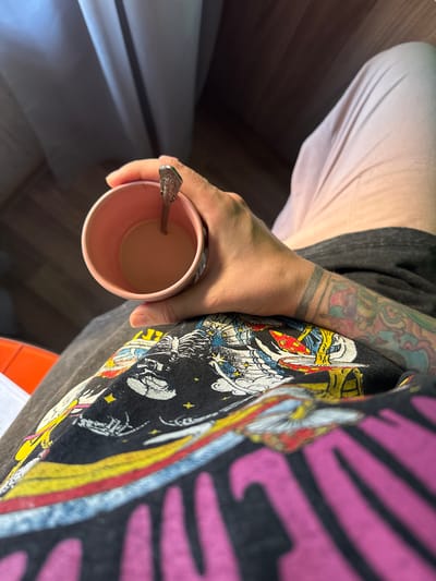 The image shows a close-up view of a person holding a pink mug filled with a light brown liquid, likely coffee or tea, with a spoon inside. The person's hand and forearm, which are covered in tattoos, are visible. They are wearing a dark t-shirt with a colorful graphic print. The background shows a blurred view of light-colored curtains and a dark, wooden floor. There are no discernible landmarks or elements that would indicate the location as Norilsk, Russia.