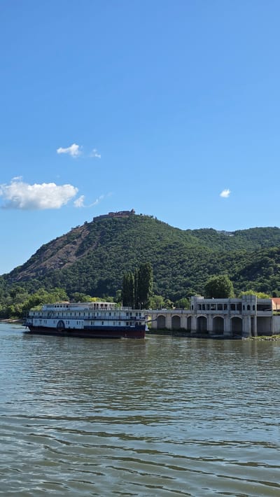 A wide river boat, painted white with dark blue trim, travels on a calm river. The boat is multi-decked with numerous windows. On the riverbank to the right, a long, arcaded building with stone columns is visible. Behind the building and the boat, a large, tree-covered hill rises steeply. At the summit of the hill, the ruins of a castle, appearing as stone structures, are clearly visible. The sky above is a bright, clear blue with a few scattered white clouds. The scene suggests a sunny day in Visegrád, Hungary, a town known for its medieval citadel overlooking the Danube River.