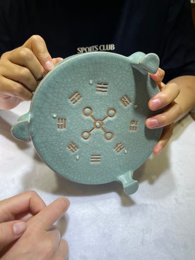 In the Futian District of China, two individuals are holding a light blue, crackled ceramic object. The object is circular with four protrusions extending outwards, resembling feet. The surface of the object is decorated with a central design of interconnected circles and lines, and around its perimeter, there are eight rectangular indentations containing Chinese characters or symbols. The object appears to be a decorative piece, possibly a lid or a small dish. One person's hand is on the left side of the object, holding it from underneath, while the other person's fingers are on the top right, interacting with the object. The background is a plain white surface, with a dark garment visible on the left, suggesting the presence of another person or clothing. The lighting appears to be indoor, with some shadows cast on the white surface. The scene conveys a sense of examination or presentation of the ceramic artifact.