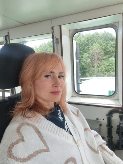 A medium shot captures a woman with shoulder-length, peachy-pink hair and bangs, wearing a white knit cardigan with a heart pattern and a dark t-shirt underneath. She is seated in what appears to be the captain's cabin of a vessel, with a black headrest visible to her left and a window with a view of trees and a white structure to her right. The interior of the cabin is visible, including a white ceiling and window frame, and some nautical equipment on the right side of the frame. The woman is looking towards the camera with a slight smile. The setting is the city of Vytegra, Russia.