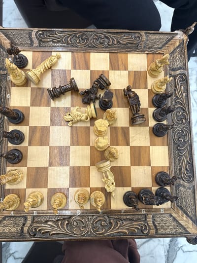 An ornate wooden chessboard, with intricately carved edges, displays an ongoing chess game. Black pieces, including rooks, knights, and pawns, are primarily positioned near the top and left of the board, some lying on their sides. White pieces, also including rooks, knights, and pawns, occupy the bottom and right. The checkered pattern of the board is visible, with light and dark squares. The setup suggests a mid-game scenario. Two people appear on the left and right side, ready to play the game.