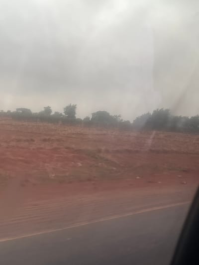 The image captures a broad, open landscape under an overcast or hazy sky, likely viewed from a moving vehicle. The dominant features include a bright, diffused sky, a line of dense, dark trees, and a vast expanse of reddish-brown earth.

The top left portion of the frame is filled with the pale, bright sky, suggesting diffused light characteristic of an overcast day. Below this, running horizontally across the middle of the frame (if rotated correctly), is a dense, irregular line of trees and shrubs, silhouetted against the bright background. Their forms are dark, making specific species difficult to discern, but they appear healthy and somewhat uniform in height.

To the right of the tree line, a wide stretch of reddish-brown, dry-looking land fills the foreground and midground. The soil appears somewhat barren or sparsely vegetated, with faint textures and subtle undulations visible on its surface. There are small, scattered darker specks on this land, possibly small plants or rocks.

The far right edge of the image features a dark, straight band, which could be the frame of a vehicle window or a section of asphalt road. Slight reflections or glare are visible on this dark surface, reinforcing the idea of the image being taken from inside a moving vehicle.

There are no people or man-made structures visible in the landscape, apart from the potential road or window frame. The scene conveys a sense of quiet, open rural space, possibly in the Makera region of Nigeria, consistent with the appearance of lateritic soils common in such areas. The time of day is daytime, but the heavy overcast conditions prevent any strong shadows or direct sunlight. No text is visible in the image.