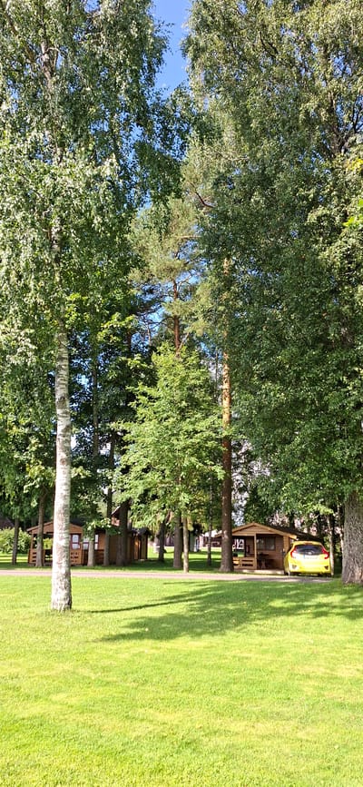 The image shows a sunny day scene in Valma, Estonia, featuring a grassy lawn, several trees, and small wooden cabins. A bright yellow car is parked in front of one of the cabins. The cabins appear to be rustic and are nestled among the trees. The overall atmosphere is peaceful and natural, suggesting a recreational area or a campground. The time of day is likely afternoon, given the long shadows cast by the trees on the lawn.