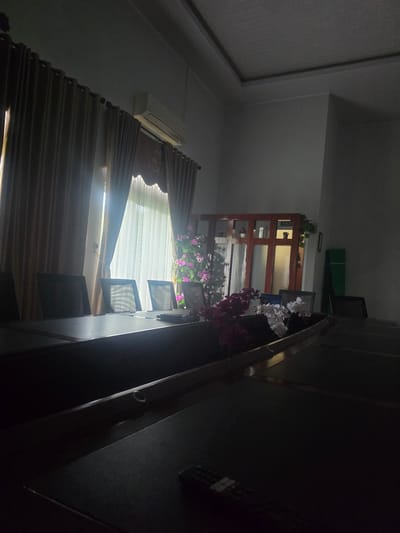 The image shows the interior of a room in Pintomakmur, Indonesia. A window with closed blinds allows light to enter. An air conditioning unit is mounted on the wall above the window. A wooden shelving unit is visible in the background, decorated with greenery and flowers. A staircase with a dark railing is positioned on the right side of the image. Several objects are scattered on the staircase.