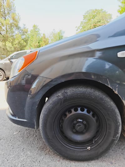 An angled, close-up shot shows the front right side of a dark gray car parked on a gravelly surface, likely in Navoiy City, Uzbekistan. The car's tire, a black EUROLUX brand, with its black rim and visible lug nuts, is prominently featured. The car's fender shows several scuff marks and some white splatters, possibly mud or paint. The headlight assembly, with its orange turn signal, is visible near the front of the car. In the background, another car, lighter in color, is partially visible, along with green trees and a bright sky, suggesting an outdoor setting. No people are present in the image.