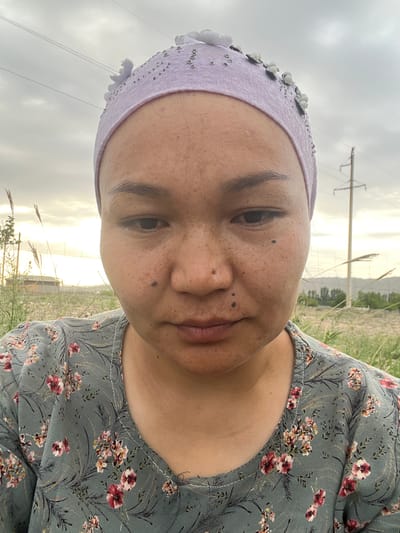 A close-up, vertically oriented, selfie of a person lying down outdoors in Samarkandek, Kyrgyzstan. The person is wearing a light purple headscarf adorned with white and silver floral embellishments. Their face is partially visible, with visible freckles and some darker marks. The person has dark eyebrows and is looking towards the camera. They are wearing a grey floral print dress. The background shows a cloudy sky with a hint of sunset or sunrise light breaking through. Power lines and a pole are visible in the distance, suggesting a rural or semi-rural setting. Tall grasses and weeds are in the foreground and surrounding the person, indicating an outdoor, natural environment. The overall mood appears casual and relaxed. The time of day is likely late afternoon or early morning due to the quality of the light. No text is visible in the image.