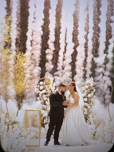 An outdoor wedding celebration is depicted in Gennevilliers, France. A man and a woman, central to the frame, stand facing each other on a white surface. The man wears a dark suit, white shirt, and has a dark beard; he holds a slender glass in his right hand. The woman wears a white strapless wedding gown and a veil, also holding a slender glass in her right hand. Their heads are tilted towards each other, suggesting an intimate interaction such as a toast or kiss.
The background is elaborate, featuring tall white floral arrangements that appear to be artificial, interspersed with golden, curvilinear structures forming an arch or decorative elements. To the left, a golden, slatted rectangular stand is visible. Numerous vertical plumes of white smoke or low-lying fog rise from the ground around the couple and decorations. Above these plumes, a series of vertical lines composed of falling bright particles or sparks are active, indicating the use of pyrotechnic effects like cold spark fountains against a darker, possibly green, backdrop. The overall scene is brightly lit.