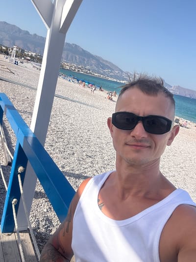 A selfie taken by a man on a sunny day at a beach in Pla Parcial Platja, Spain. The man is wearing a white tank top and black sunglasses. He has short, spiky hair and a tattoo on his arm. The beach is made of pebbles, and there are lounge chairs and umbrellas set up. In the background, there are mountains and the sea. The man is standing on a wooden deck with a blue railing.