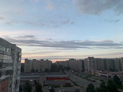 The image depicts a panoramic view of an urban environment, likely Severodvinsk, Russia, during the twilight hours. In the foreground, a multi-story apartment building with a light-colored facade dominates the left side of the frame, showcasing numerous windows and balconies. Across the street, several other residential buildings of varying heights and architectural styles are visible, forming a dense urban landscape. A main road runs through the scene, with cars parked along its sides and a few in motion. A low-rise commercial building with a red-and-white facade is situated between the apartment complexes and the road. Trees are interspersed throughout the urban setting, particularly in the mid-ground and foreground. The sky above is a gradient of pale blue and gray, with scattered clouds indicating the transition from day to night. Two thin, dark power lines traverse the sky diagonally from the upper left to the upper right of the image.
