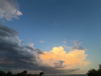 The image captures a dramatic sky with a mix of dark, ominous clouds and brightly illuminated, golden cumulus clouds. The lower portion of the frame shows a silhouetted treeline and the rooftops of buildings, suggesting a semi-urban or suburban environment. The light suggests it's either sunrise or sunset, with the sun's rays catching the edges of the lighter clouds. There are no people or active subjects visible in the scene. The environment appears to be outdoors, likely a residential area in Slobozia, Moldova.
