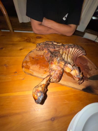 A cooked lamb carcass sits on a wooden cutting board. The lamb appears heavily seasoned and roasted, with the rib cage and leg bones exposed. The board rests on a wooden table, accompanied by a plate and a person in a black t-shirt. The scene is indoors, potentially a home dining setting.