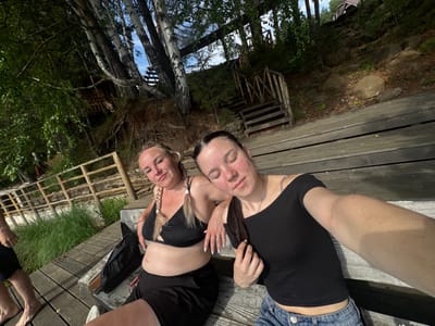 The image shows two young women sitting on a wooden deck outdoors. The woman on the left has blonde hair in pigtails and is wearing a black bikini top and black shorts. She is leaning back and smiling. The woman on the right has dark hair and is wearing a black off-the-shoulder top and denim shorts. She is leaning her head on the other woman's shoulder and has her eyes closed. They appear to be taking a selfie. The background shows trees, a wooden fence, and a wooden staircase leading up a hill. The scene appears to be in a natural setting, possibly a lake or riverfront, with a rustic aesthetic. The location of Pozdyshki, Russia is provided as context for the image.